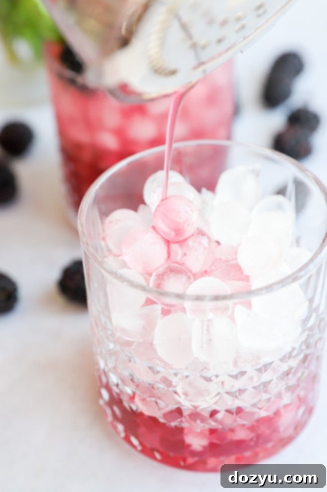 Image of the blackberry gin mixture being poured from a shaker into a glass filled with ice, demonstrating the straining step of cocktail preparation.