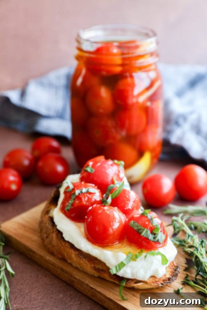 A slice of toasted bread topped with creamy cheese, cherry tomato confit, and fresh basil sits on a wooden board. In the background, a jar filled with cherry tomatoes and a few loose tomatoes are visible.