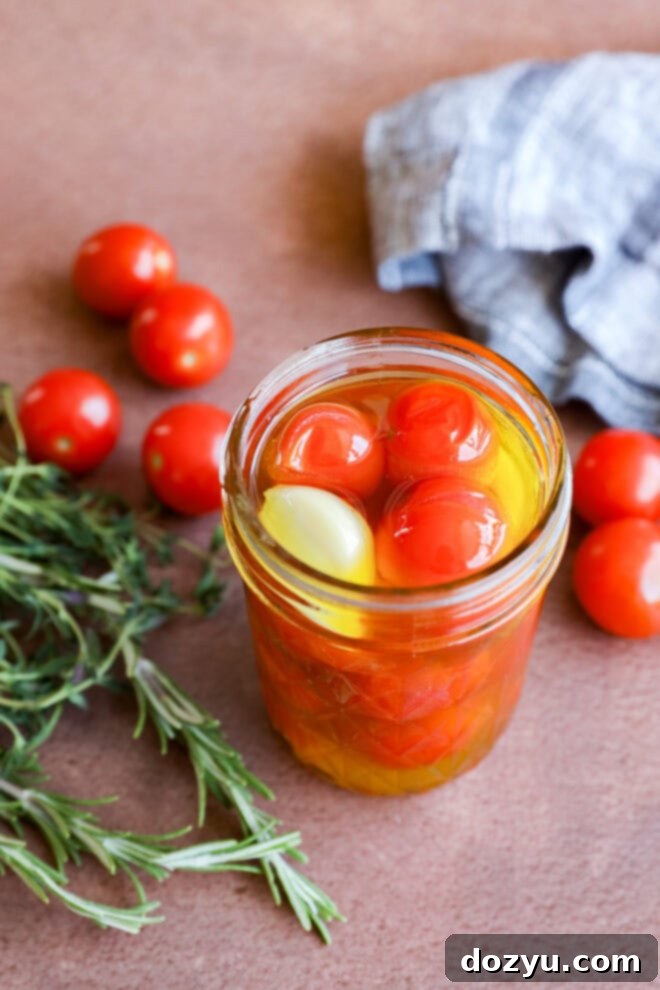 A jar of cherry tomato confit—cherry tomatoes, olive oil, and a clove of garlic—rests on a brown surface beside fresh rosemary, loose cherry tomatoes, and a blue checkered cloth.