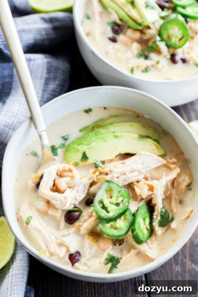 Dreamy Slow Cooker White Chicken Chili 2 A steaming bowl of creamy white chicken chili garnished with fresh cilantro, avocado, and lime wedges, with a spoon ready for enjoyment.