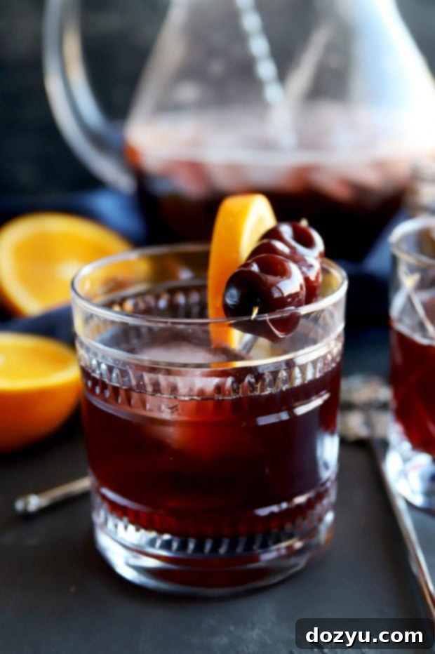 A hand pouring Perfect Manhattan Punch from a large glass pitcher into a cocktail glass filled with ice, ready to serve