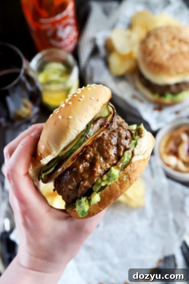 A hand holding a gourmet Miso Glazed Sriracha Burger, highlighting its impressive height and delicious layers.