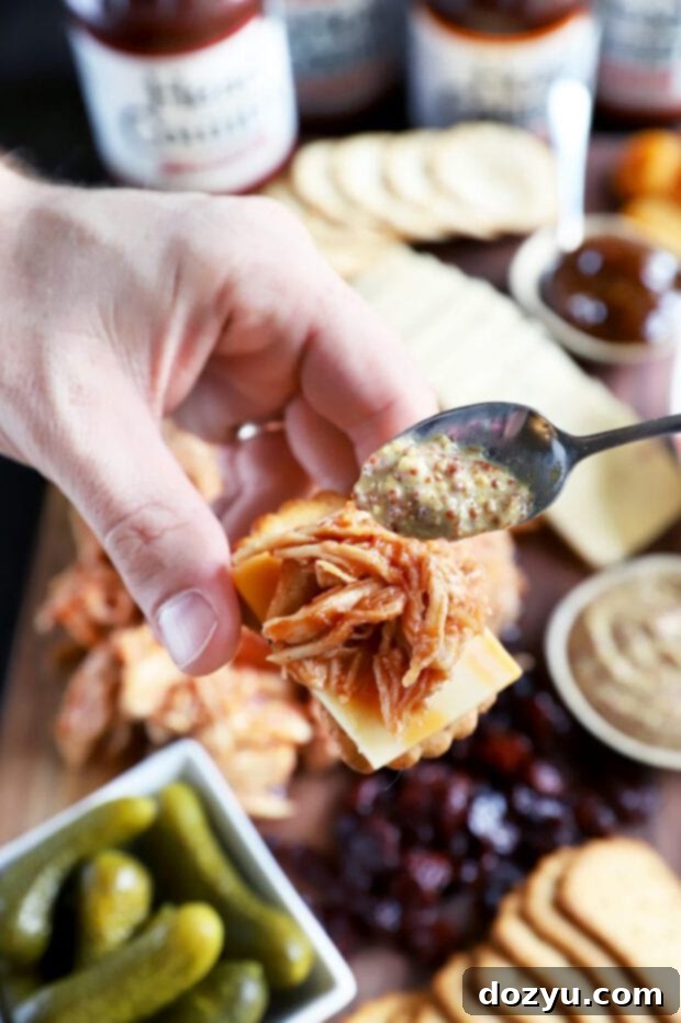 A person spooning whole grain mustard over a cracker topped with BBQ meat and cheese.