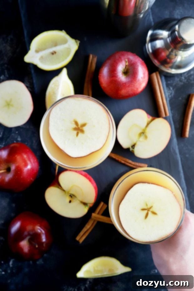 Overhead photo of two golden whiskey apple cocktails garnished with apple slices in elegant coupe glasses, set against an autumn-themed background.