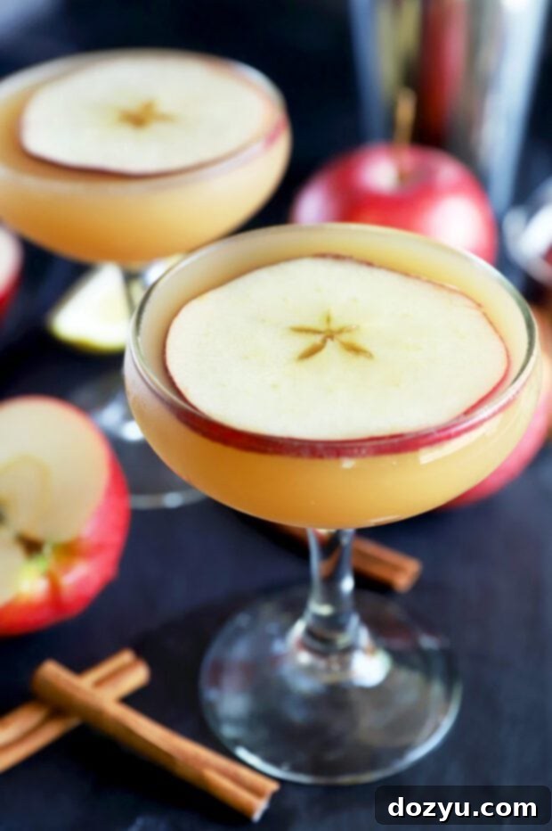 Close-up side view of an Apple Whiskey Sidecar cocktail in a coupe glass, with blurred apples in the background, highlighting the drink's golden hue and garnish.