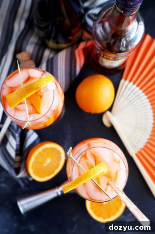 Two perfectly prepared Aperol Spritz cocktails with ice and orange garnishes on a table, surrounded by an orange, sliced oranges, a bottle, a jigger, and a fan, evoking a warm, inviting atmosphere.
