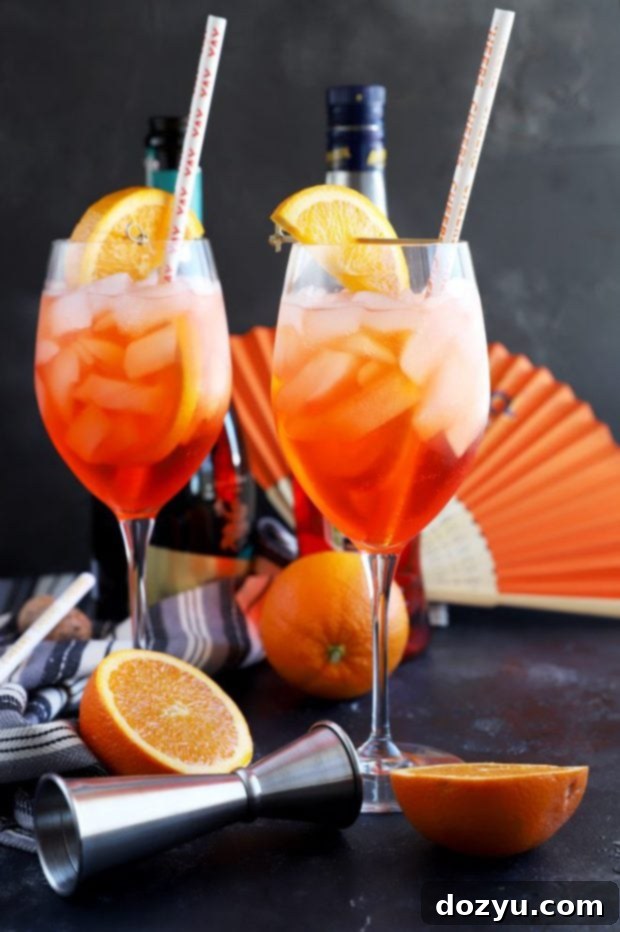 A hand holds a tall glass filled with a bright orange Aperol Spritz, ice, and an orange slice, with a straw, suggesting a refreshing drink on a warm day.