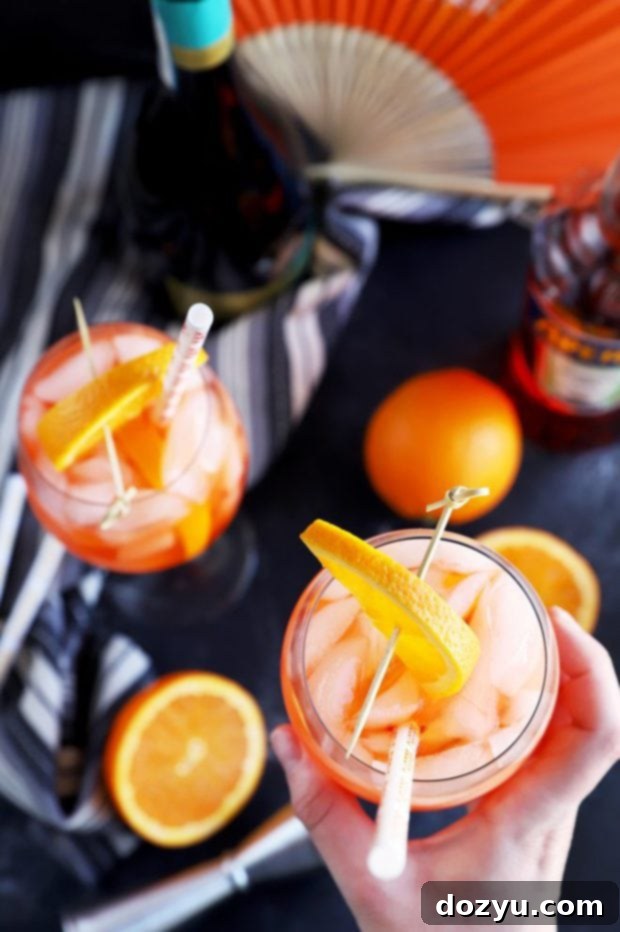A vibrant Aperol Spritz cocktail, featuring ice and a fresh orange slice, held in a hand against a blurry background, emphasizing its refreshing appeal.