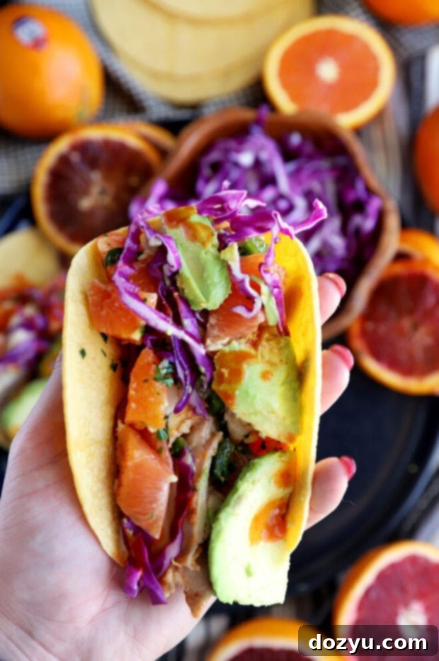 A hand holds a single Blood Orange Honey Ginger Chicken Taco, showcasing the roasted chicken, fresh salsa, and vibrant toppings nestled within a warm tortilla, ready for a bite.