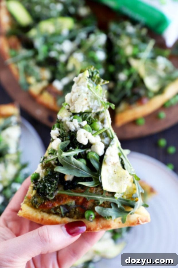 A hand holding a slice of Pesto Green Pizza, showing the delicious layers and readiness to eat.
