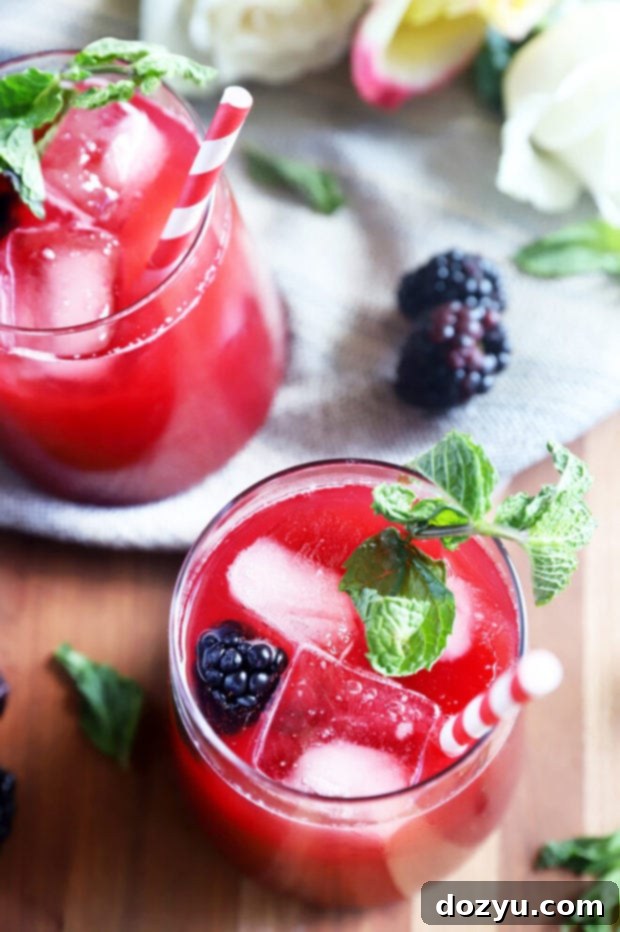 A tall glass of Blackberry Vodka Limeade garnished with fresh blackberries and mint, with a vibrant purple hue against a bright background.