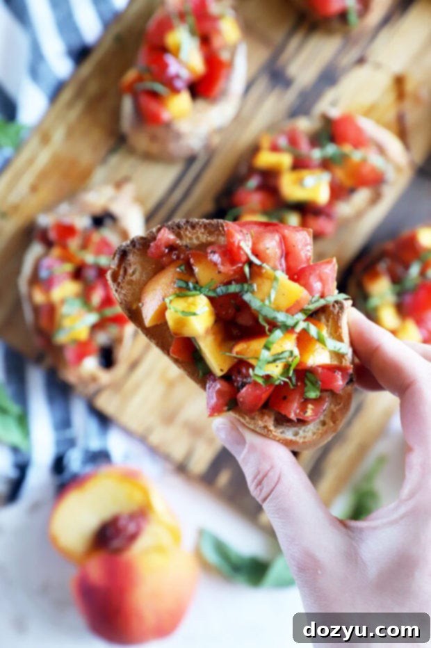 A hand holds a single piece of summer crostini, featuring a colorful topping of tomato and peach.