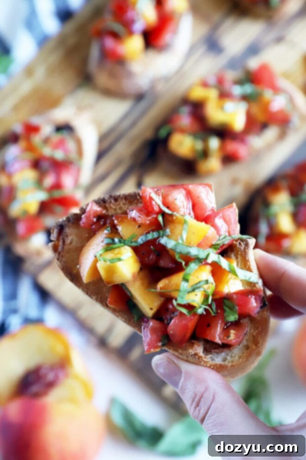 Close-up image of a hand holding a single piece of Tomato Peach Bruschetta, ready to eat.