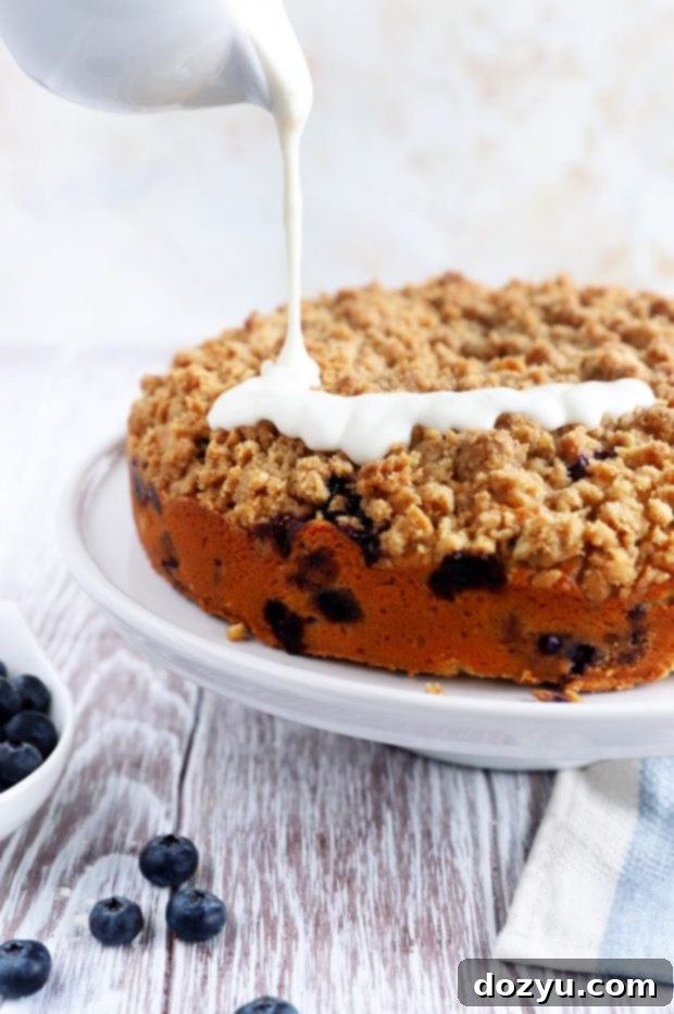 Lemon cream cheese glaze being drizzled generously over a warm Blueberry Lemon Streusel Coffee Cake on a wire rack, enhancing its visual appeal.