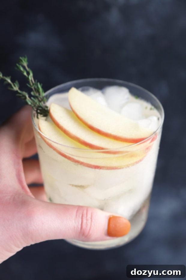 A hand holding a perfectly garnished Thyme Apple Gin and Tonic, showcasing the drink's autumn aesthetic