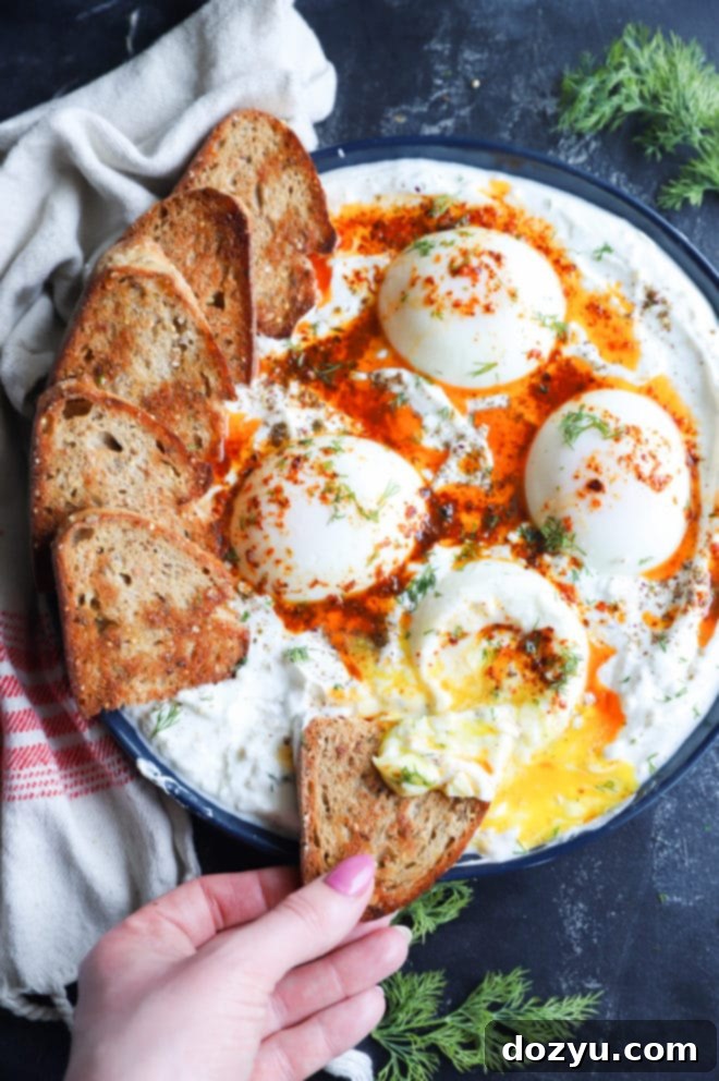 A hand holding a piece of toasted bread, dipping it into the delicious Turkish Eggs, showcasing the rich textures and vibrant colors.