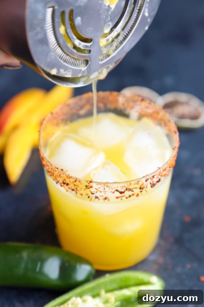 Straining spicy mango margarita into a rimmed glass with ice and mango garnish
