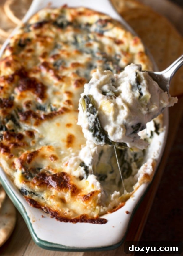 A close-up of a bubbling Spinach and Artichoke Goat Cheese Dip in a baking dish, garnished with fresh herbs.