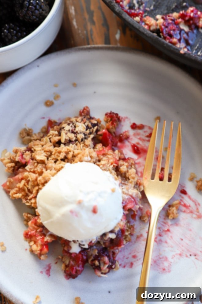 Overhead photo of a serving of apple blackberry crumble on a white plate, accompanied by a fork and a scoop of ice cream