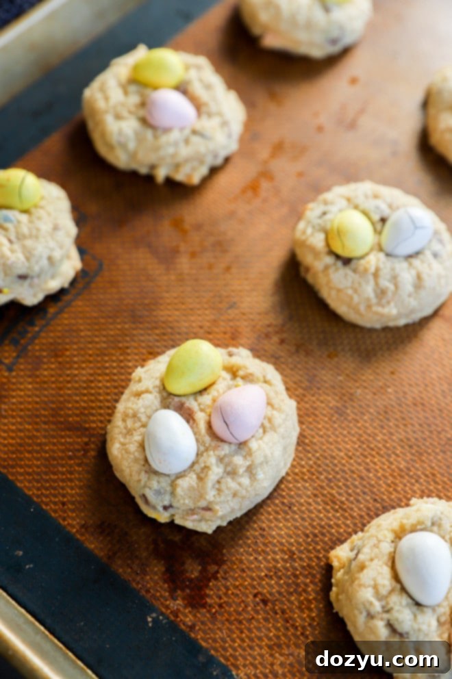 Freshly baked Easter egg cookies, adorned with colorful Mini Eggs, cooling on a baking sheet with a silicone mat.