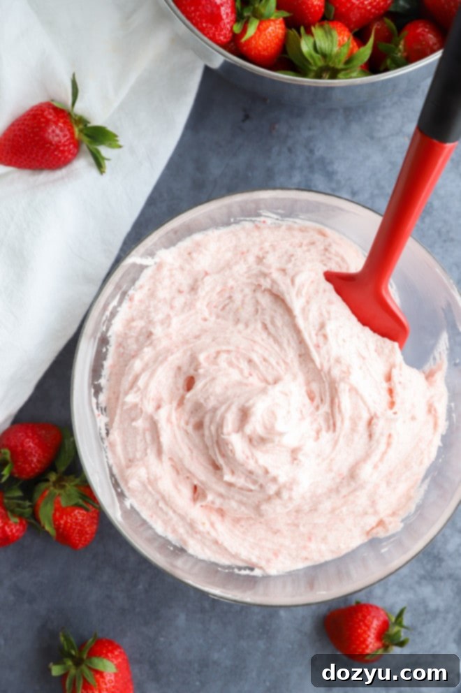 Strawberry buttercream frosting in a bowl with a spatula and fresh fruit, ready to be spread onto a delicious dessert