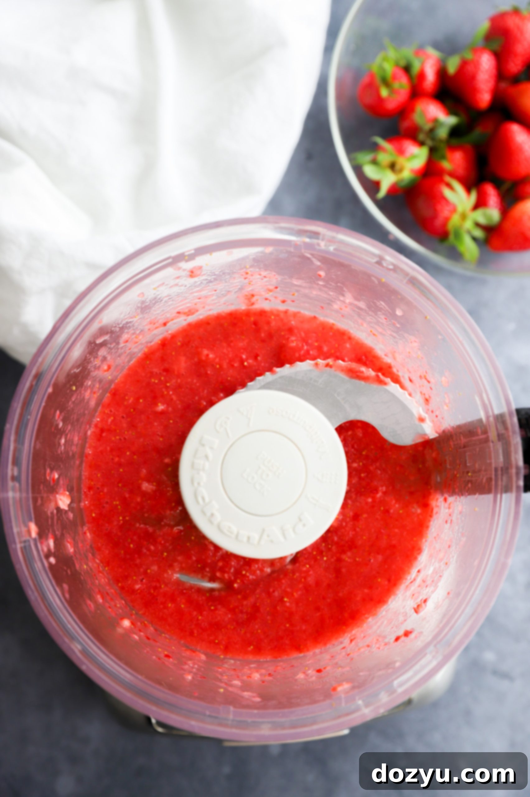 Strawberry puree in a food processor with fresh berries in a bowl, showing the initial step of preparing the fruit for frosting