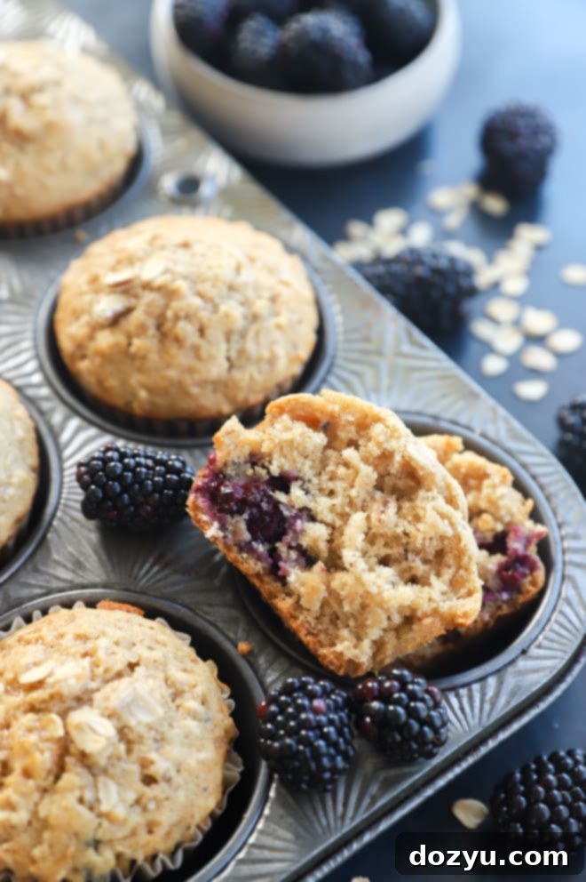 A Honey Blackberry Oatmeal Muffin cut in half, revealing its tender interior and juicy blackberries, placed in a muffin tin with other whole muffins.