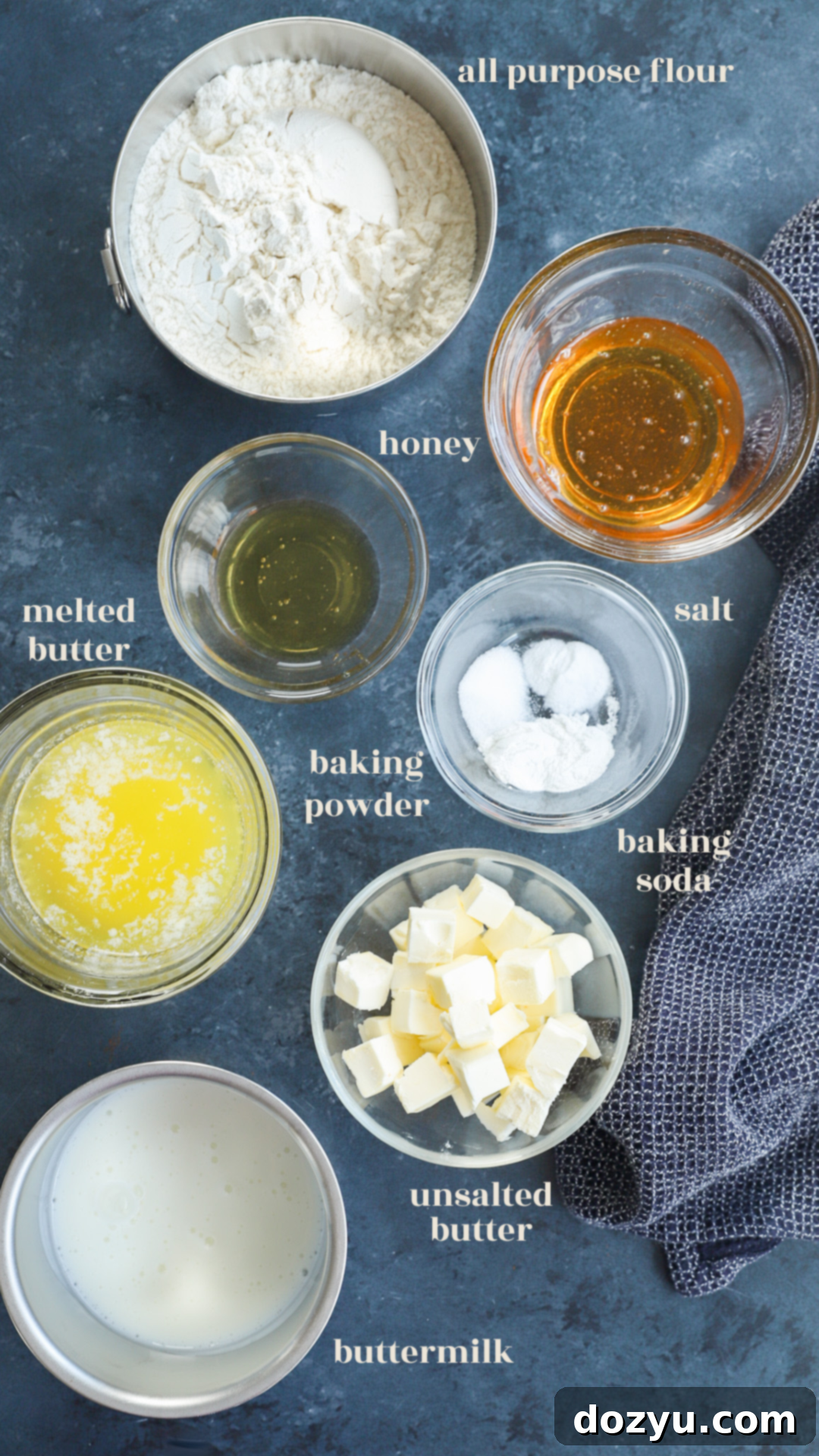 A flat lay image displaying all the essential ingredients for baking honey butter biscuits: flour, butter, honey, buttermilk, salt, baking powder, and baking soda.