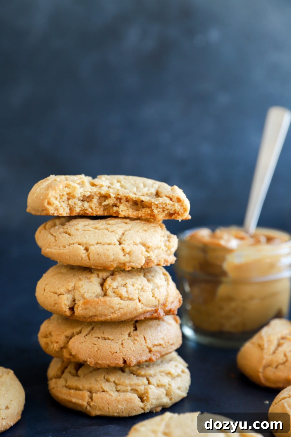 Irresistibly Chewy Peanut Butter Cookies 11 A beautiful arrangement of chewy peanut butter cookies piled on a white plate, with a wooden spoon and a jar of peanut butter in the background.