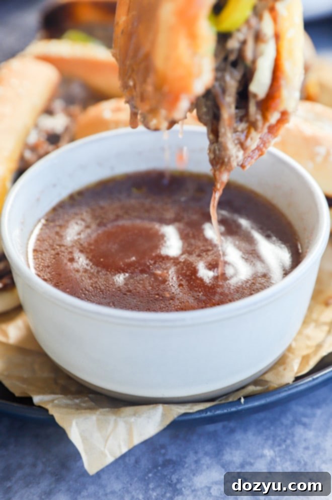 A French dip sandwich being dipped into a small, elegant white bowl filled with golden au jus, emphasizing the dipping experience.