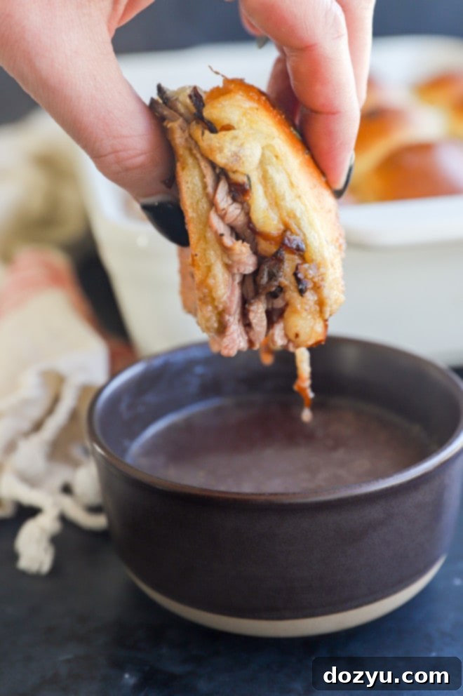 A mini roast beef slider being dipped into a small white bowl of au jus, with a full slider on a cutting board in the background.