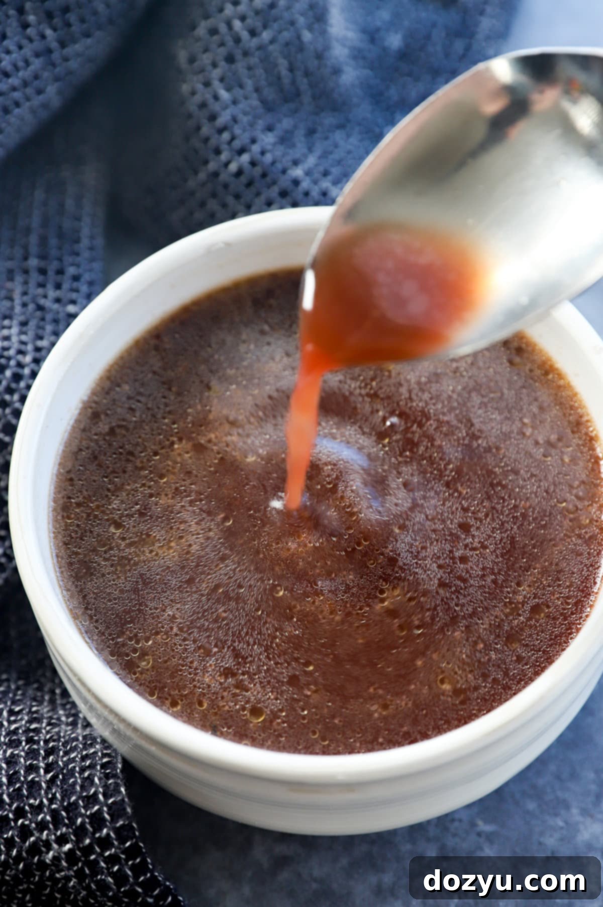 A hand using a ladle to scoop golden au jus into a small white bowl, showing the smooth consistency of the sauce.