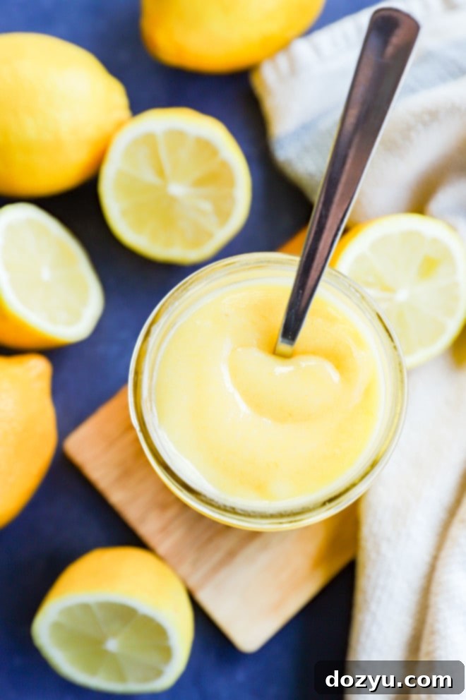 Effortless Zesty Lemon Curd 2 A mason jar filled with creamy yellow lemon curd, surrounded by fresh whole and sliced lemons, with a spoon resting in the curd.