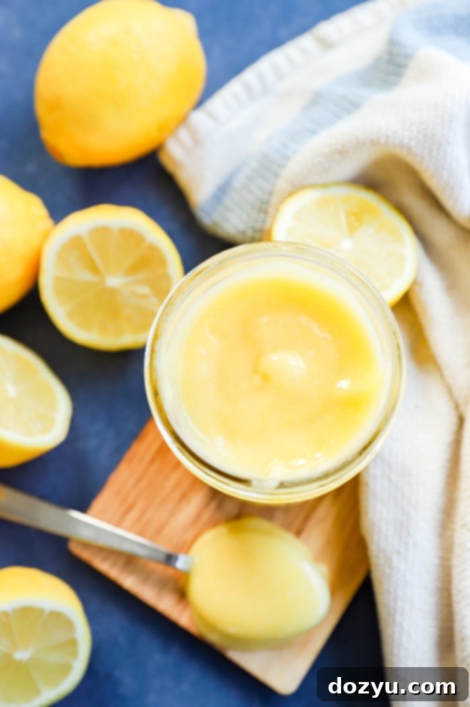 Effortless Zesty Lemon Curd 6 A glass jar filled with lemon curd, a spoon resting in it, surrounded by fresh lemons and a white linen towel, emphasizing freshness and homemade quality.