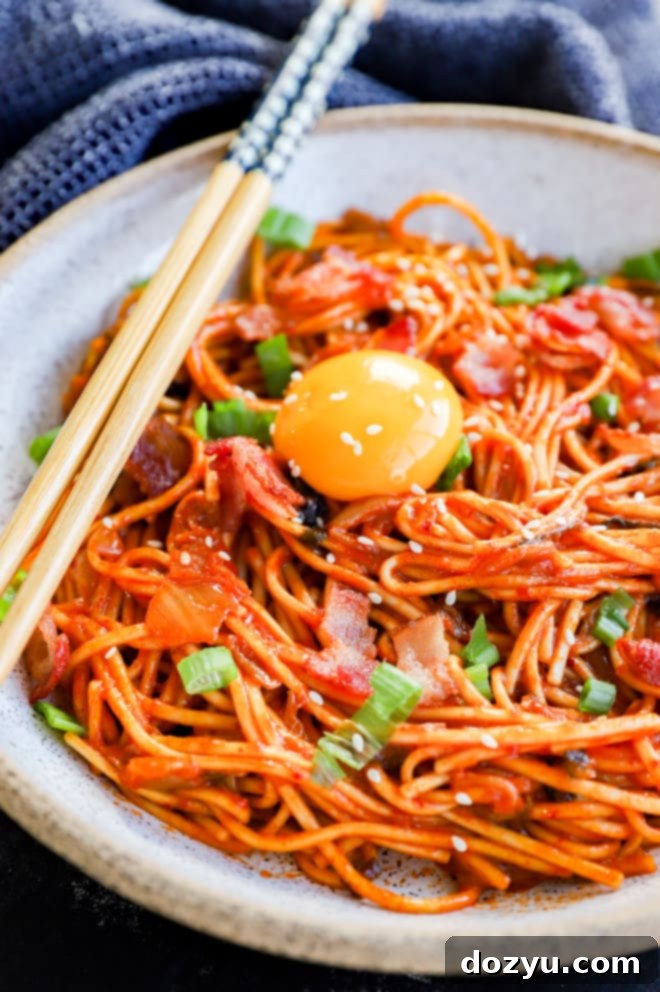 A vibrant bowl of creamy Kimchi Udon noodles with chopsticks, garnished with fresh green onion and an egg yolk