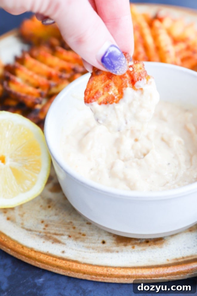 Close-up of a hand dipping a crispy french fry into a bowl of savory roasted garlic aioli.