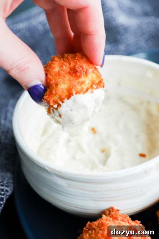 A hand holding a perfectly fried, panko-crusted potato cheese ball, with a dollop of creamy aioli on top, showcasing its delicious appeal.