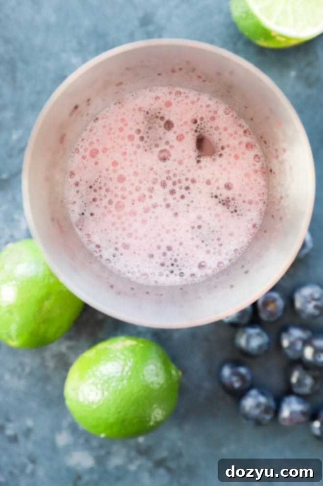 A cocktail shaker being vigorously shaken to mix a blueberry margarita, with ice visible inside.