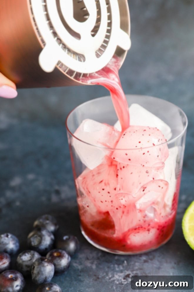 A blueberry margarita being strained from a cocktail shaker into a glass filled with ice and garnishes.
