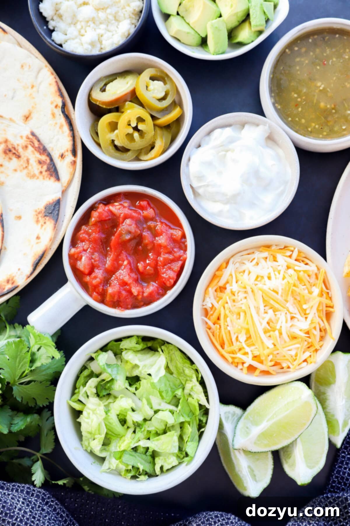 Image of a variety of toppings including shredded lettuce, lime wedges, shredded cheese, sour cream, salsa verde, salsa, pickled jalapeno, and tortillas