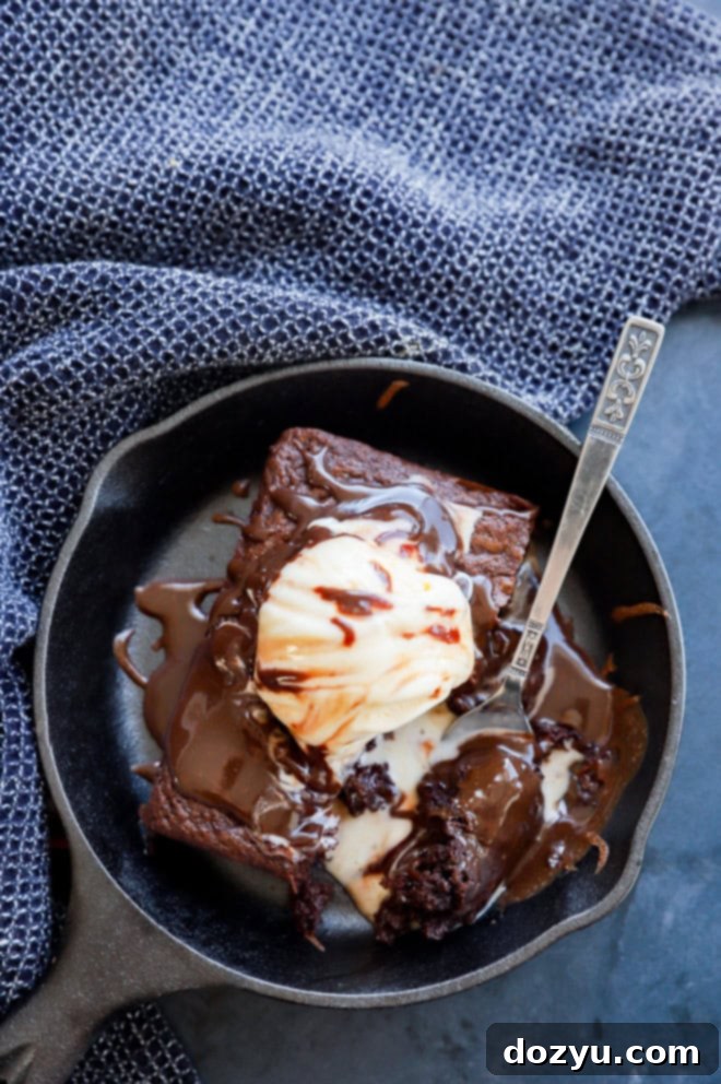A fork taking a delicious bite out of a warm chocolate brownie served in a hot skillet, topped with melting ice cream.
