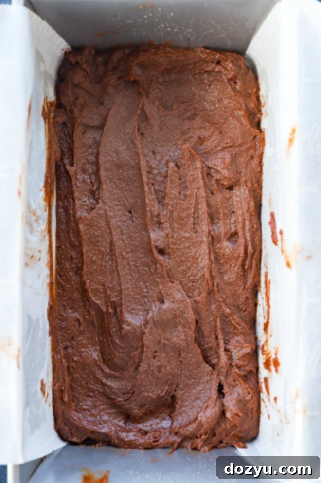 Rich brownie batter with chocolate chips poured into a parchment-lined loaf pan, ready for baking.