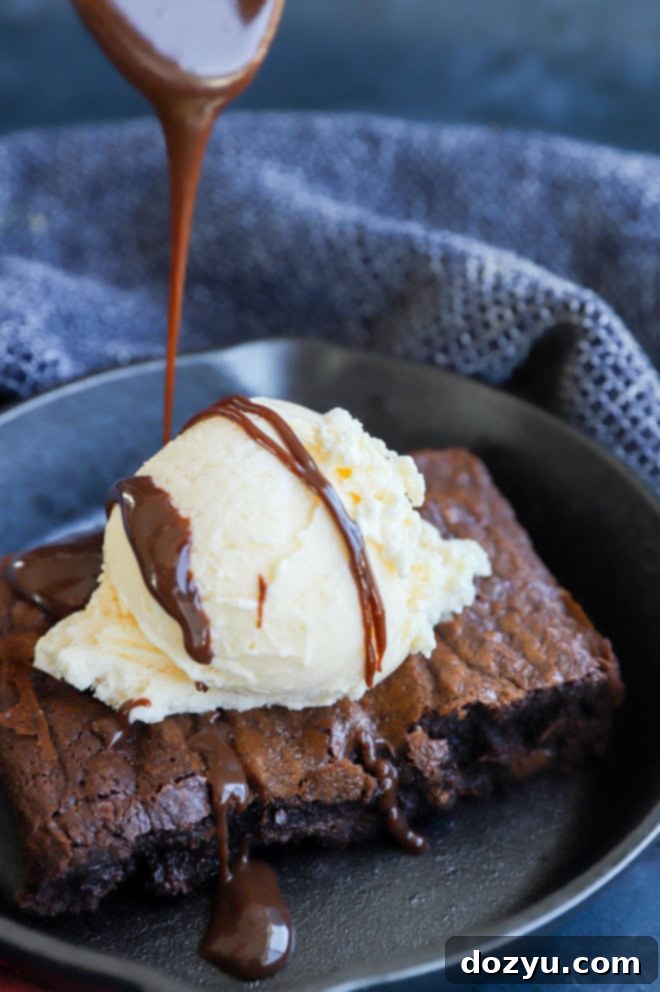 A hand drizzling rich hot fudge sauce over a scoop of vanilla ice cream on a warm brownie in a sizzling skillet.