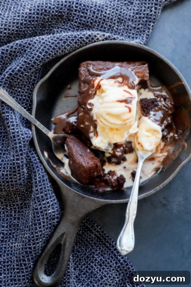 A sizzling brownie sundae in a hot skillet, garnished with melting ice cream, hot fudge sauce, and two forks, ready to be shared.