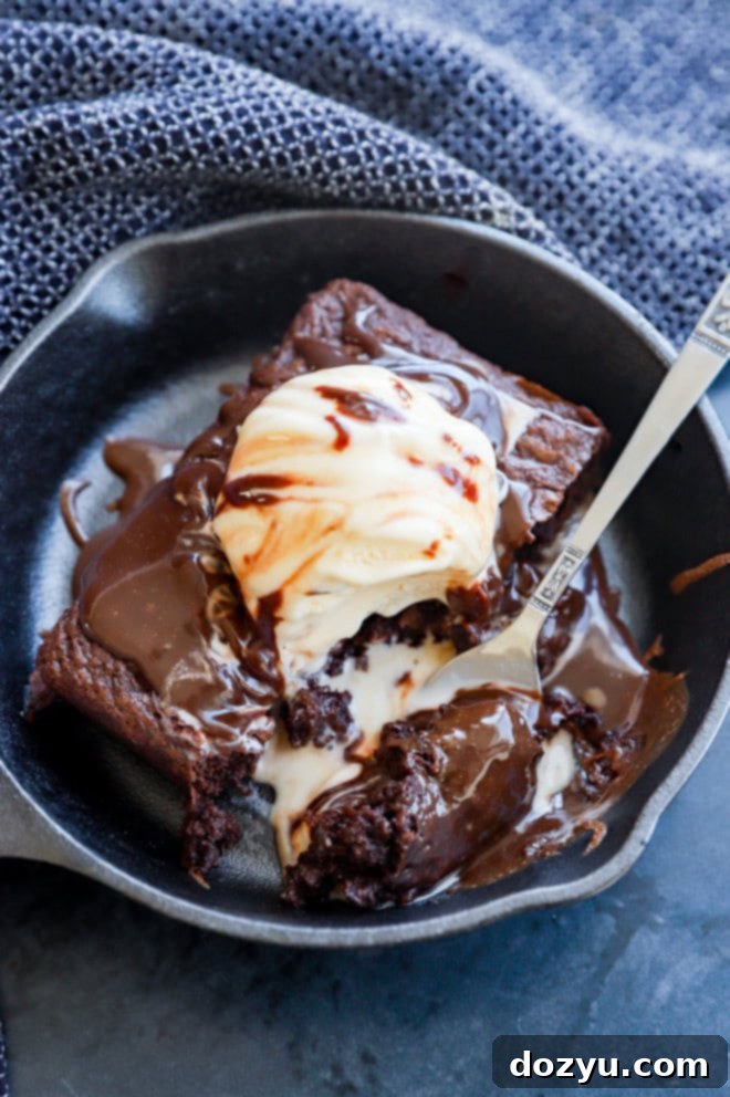 A fork lifts a bite of rich, fudgy brownie with hot fudge sauce from a hot cast iron skillet.