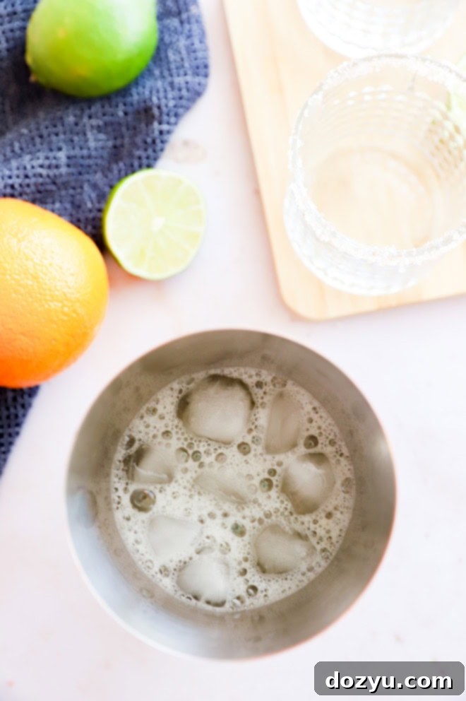 Cocktail shaker filled with ice and a Patron margarita mix, with lime slices on the side