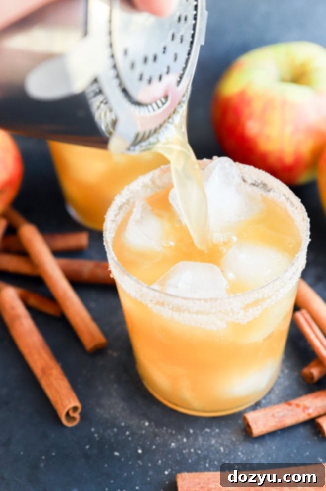 Pouring a freshly mixed apple cider margarita into a cinnamon sugar rimmed glass filled with ice, illustrating the final step.