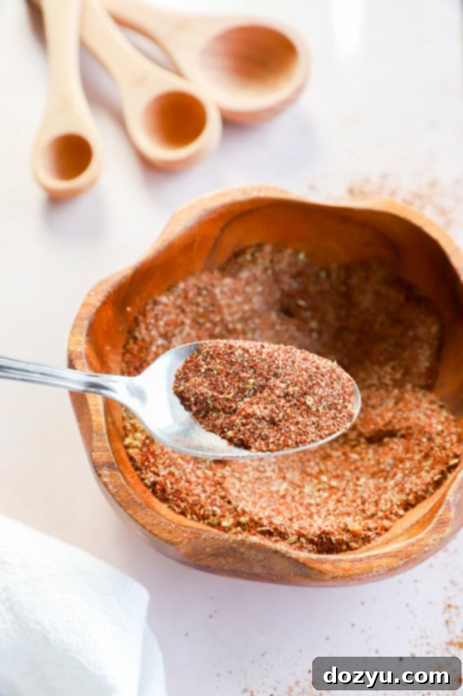 Make Your Own Chili Seasoning Mix 4 A silver spoon scoops a generous amount of homemade chili seasoning from a bowl, with blurred measuring spoons and spices in the background, emphasizing the blend's texture and richness.