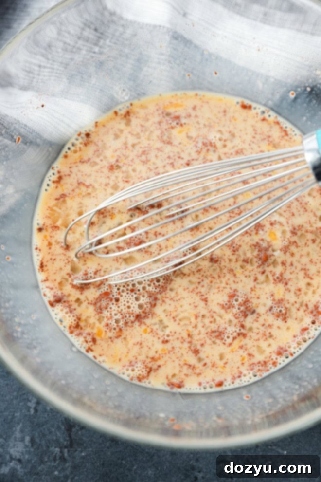 A close-up shot of a creamy, light brown cinnamon custard mixture in a glass bowl, with a metal whisk resting in it, ready for French toast preparation.