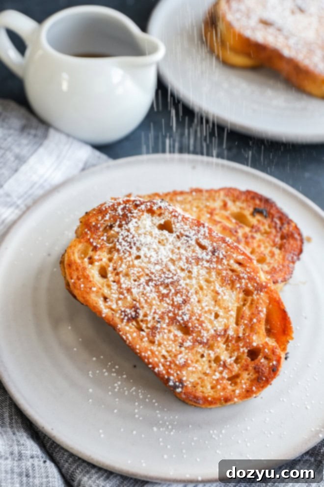 A hand sprinkles a fine dusting of powdered sugar over golden-brown slices of sourdough French toast on a white plate, ready to be served.